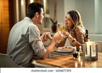 Happy woman and her boyfriend holding hands while toasting with Champagne while having dinner at dining table. 