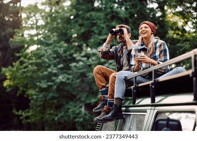 Happy woman and her boyfriend using binoculars while sitting on roof of their camper trailer and spending a day in the woods. Copy space.