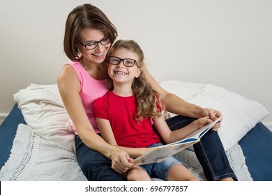 Happy Woman With Her  Daughter Child,  Reading Together A Book At Home. Mom And Daughter Wearing Glasses.