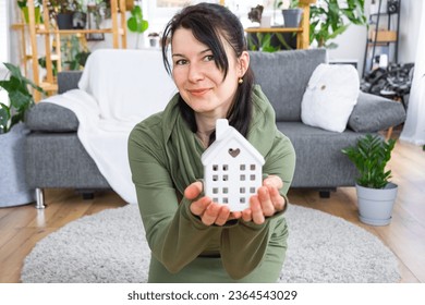 A happy woman in her house holds in her hands a miniature figure of a house and key in the interior. Dream house project, real estate purchase, insurance, mortgage, rent, reservation - Powered by Shutterstock