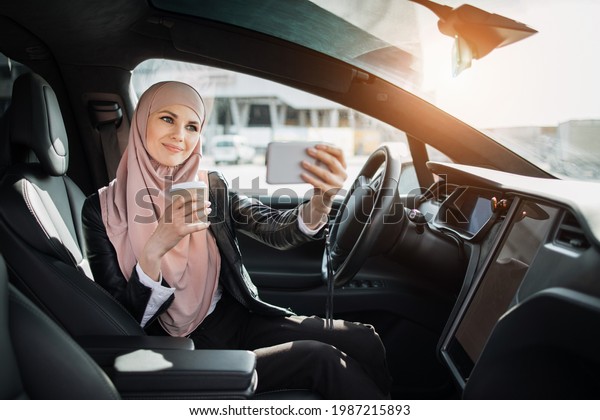 Happy woman in hijab drinking\
fresh coffee and talking during video call on smartphone. Business\
lady sitting inside modern electric car during online\
conversation.