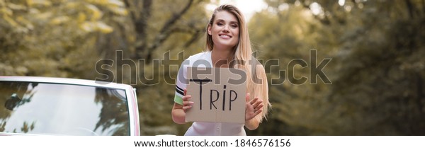 happy woman holding card with trip lettering near\
cabriolet, banner
