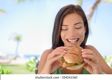 Happy Woman Holding Eating Fresh Burger Sandwich At Beach. Young Female Is Sitting At Outdoor Restaurant. Beautiful Tourist Is Having Fresh Burger During Summer Vacation Holidays.