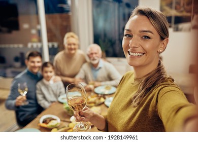 Happy woman holding glass of wine and taking selfie with her extended family during a meal at home.