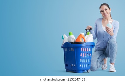 Happy Woman Holding A Liquid Soap Bottle And Full Shopping Basket, She Is Buying Cleaning Products At The Store