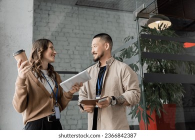 happy woman holding paper cup and tablet while chatting with asian colleague during coffee break - Powered by Shutterstock