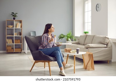 Happy Woman At Home Enjoys Her Takeout Lunch Delivered By Food Delivery Service. Young Woman With Container Of Salad In Her Hands Sits In Armchair Near Paper Bag And At Table On Which Food.