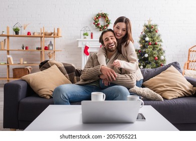 happy woman hugging husband and pointing at laptop on sofa in living room - Powered by Shutterstock