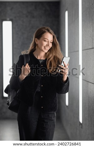 happy woman in jacket holding backpack and using smartphone near lighting of fluorescent lamps