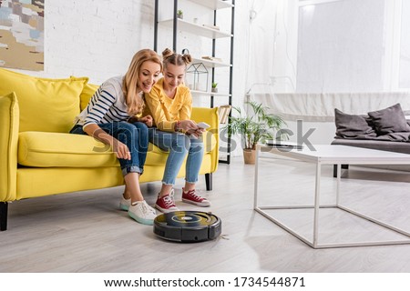 Happy woman and kid with smartphone smiling and looking at robotic vacuum cleaner near coffee table on floor in living room Stock photo © 