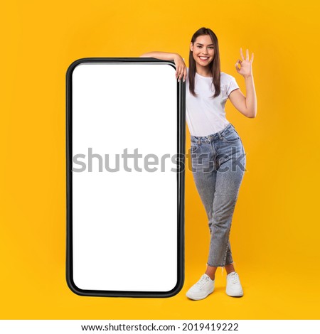 Happy Woman Leaning On Big Smartphone With Blank White Screen And Gesturing Ok Sign, Cheerful Lady Recommending New App Or Website, Standing On Yellow Background, Mock Up Image, Full Body Length