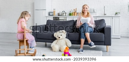 happy woman listening music on sofa near daughter riding rocking horse, banner