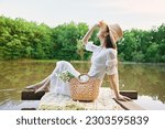 happy woman in a long light dress is resting by the lake sitting on a plaid and enjoying the view