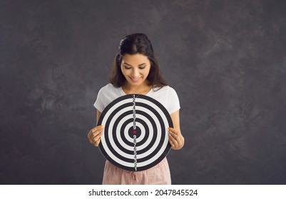 Happy Woman Looking At Dartboard Target Which She Holds In Her Hands While Standing On Gray Background. Concept Of Achieving Goals, Success, Defining Business Goals And Finding Target Audience.