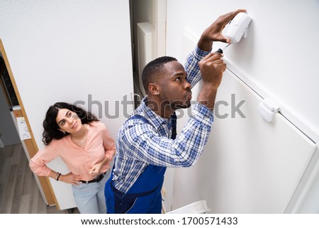 Happy Woman Looking At Electrician Installing Security System Motion Detector On Wall