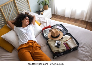 Mujer feliz tumbada en la cama al lado de una maleta abierta llena de ropa, pasaporte y sombrero, lista para ir de vacaciones.