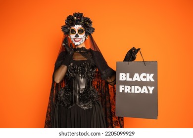 happy woman in mexican day of dead makeup and costume holding black friday shopping bag isolated on orange