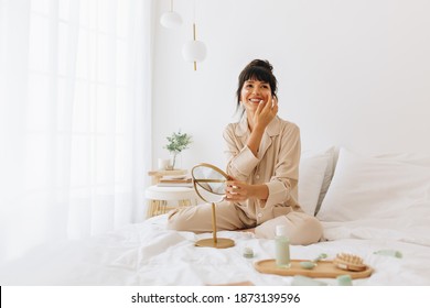 Happy Woman In Night Suit Sitting On Bed And Applying Face Cream. Woman Using Beauty Products At Home.