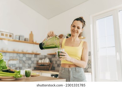 Happy woman on detox diet, pouring green cocktail from mixer into glass in kitchen - Powered by Shutterstock