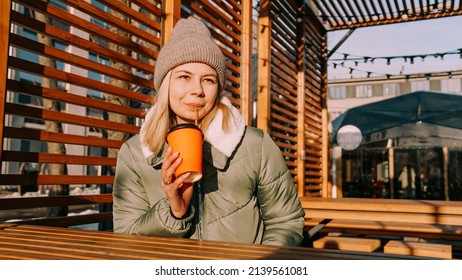 Happy Woman On A Winter Sunny Day At A Street Food Fair In Space For Relaxing And Meeting Friends. She Drinks Coffee Or Mulled Wine From An Orange Cup