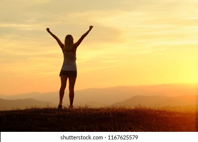 Happy woman with open arms stay on the peak of the mountain cliff edge under sunset light sky enjoying the success, freedom and bright future. - Shutterstock ID 1167625579