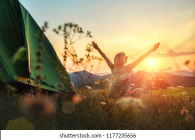 Happy woman with open arms stay near tent around mountains under sunset light sky enjoying the  leisure and freedom.