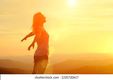 Happy woman with open arms stay on the peak of the mountain cliff edge under sunset light sky enjoying the success, freedom and bright future.