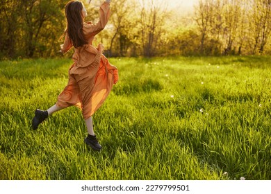 a happy woman in an orange dress illuminated by the rays of the setting sun happily jumps in a green field in the park, enjoying nature and a warm summer day. Horizontal photo - Shutterstock ID 2279799501