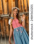 Happy woman in a pink dirndl with a blue apron and a flower crown smiles and waves, enjoying a sunny day outdoors in front of a rustic wooden backdrop at oktoberfest or beer garden in munich 