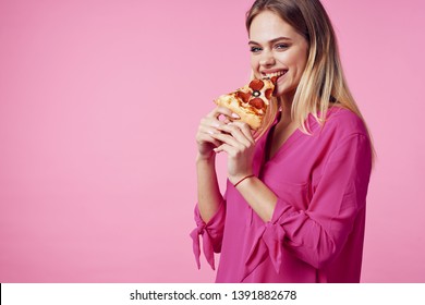 A happy woman in a pink shirt is eating a slice of pizza and smiling
