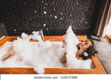 Happy woman playing with foam in big bath with wooden edge. Relaxation in spa
