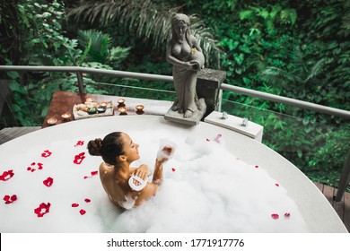 Happy Woman Playing With Foam In Big Bath Tub With Bubbles And Rose Flowers Petals. Relaxation In Spa. Beauty Treatment.