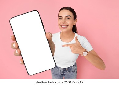 Happy woman pointing at mobile phone with blank screen on pink background. Mockup for design