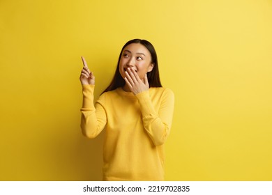 Happy Woman Pointing Up. Amused Asian Girl Points Fingers Away Closed Mouth With Hand At The Empty Copy Space. Pretty Female Smiling, Isolated On Yellow. Advertising Concept 