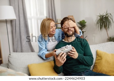 Happy Woman Presenting Gift To Husband. Excited Young Woman Closed Eyes Presenting Good Unexpected Present To Husband At Home. Loving Girlfriend Making Romantic Surprise To Boyfriend 