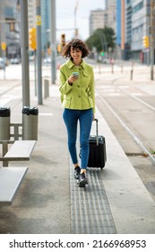 Happy Woman Pulling Rolling Suitcase And Text Messaging Using Smart Phone Outdoors In City