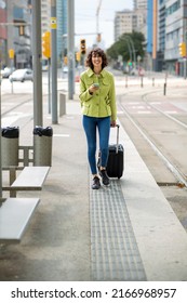 Happy Woman Pulling Rolling Suitcase And Text Messaging Using Mobile Phone Outdoors In City