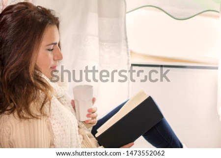 Happy woman reading a book while drinking a hot coffee at the edge of the window, in autumn.