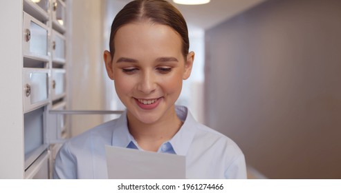 Happy Woman Reading Received Letter Standing Among Mailboxes. Close Up Portrait Of Smiling Young Female Opening Letterbox And Receiving Good News In Apartment House Lobby