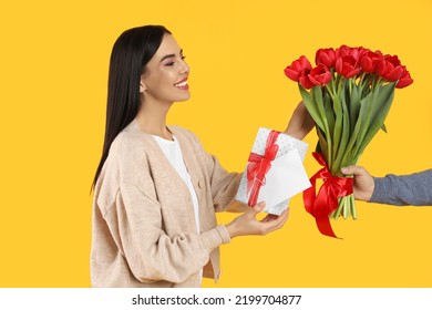 Happy Woman Receiving Red Tulip Bouquet, Greeting Card And Gift Box From Man On Yellow Background. 8th Of March Celebration