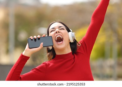 Happy woman in red holding phone as a mic singing in the street listening to music with headphones - Shutterstock ID 2195125353
