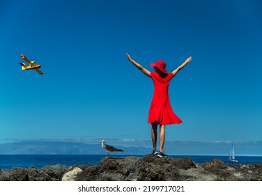 Happy Woman In Red Standing On The Rocky Ocean Shore With Open Hands. Calm Ocean Water And Pure Blue Sky At The Background.