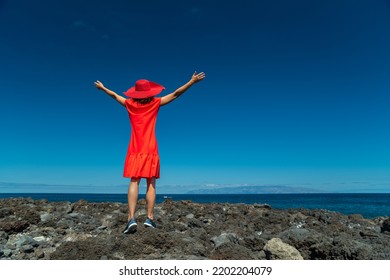 Happy Woman In Red Standing On The Rocky Ocean Shore With Open Hands. Calm Ocean Water And Pure Blue Sky At The Background.