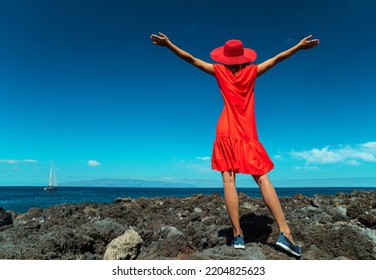 Happy Woman In Red Standing On The Rocky Ocean Shore With Open Hands. Calm Ocean Water And Pure Blue Sky At The Background.