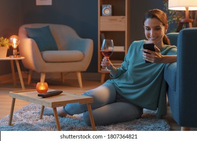 Happy Woman Relaxing At Home, She Is Drinking A Glass Of Wine And Connecting With Her Smartphone