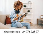 Happy Woman Relaxing at Home, Smiling and Holding Tablet PC while Listening to Music with Headphones on Cozy Couch