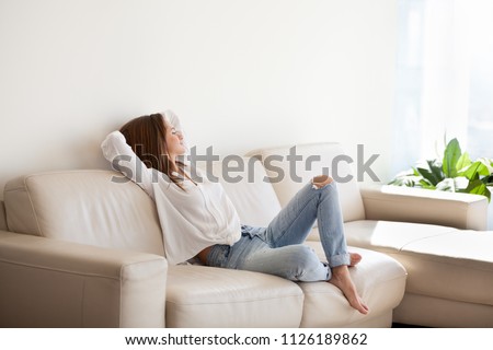 Happy woman relaxing on comfortable soft sofa enjoying stress free weekend at home, calm satisfied girl stretching on couch thinking of pleasant lazy day, dreaming and planning sunday morning