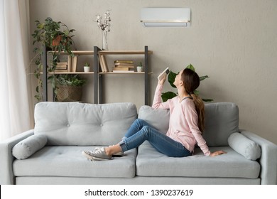 Happy Woman Rests On Couch In Modern Cozy Living Room Looks Up On Wall Holds Cooler System Remote Controller Air Conditioner User Enjoy Fresh Air In Hot Summer Day, Set Comfortable Temperature Indoors