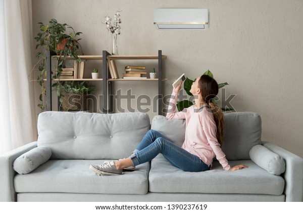 Happy woman rests on couch in modern cozy living\
room looks up on wall holds cooler system remote controller air\
conditioner user enjoy fresh air in hot summer day, set comfortable\
temperature indoors