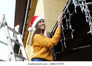 Happy woman in Santa hat decorating house with Christmas lights outdoors - Powered by Shutterstock